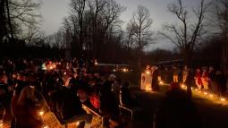 A large group sits on benches at night surrounded by lanterns as speakers read from a podium